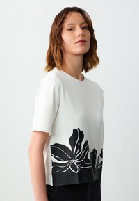 Weißes Kurzarmoberteil mit einem schwarzen Blumenmuster am Saum. Glatter Stoff mit einer lockeren Passform und rundem Halsausschnitt.