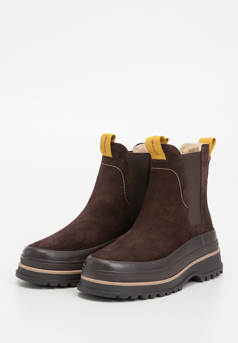 Bruna mocka anklar stövlar med en kraftig gummisula. Har elastiska sidopaneler och en gul dragflik för enkel påtagning. Texturerad yta.