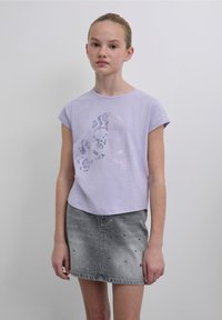 T-shirt lilla a maniche corte con grafica lucida di teschio e ossa incrociate. Abbinata a una gonna in denim con borchie argentate e orlo sfrangiato.