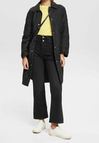Cappotto lungo trapuntato nero, t-shirt gialla, jeans neri a vita alta e sneaker bianche. Outfit casual con un design elegante e una vestibilità strutturata.