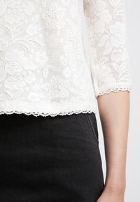 Top in pizzo bianco con motivi floreali, maniche a tre quarti e orlo scallop, indossato sopra pantaloni in tessuto scuro.