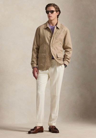 Young man wearing beige blazer, lavender sweater, cream trousers, brown tassel loafers, and sunglasses standing against plain background.