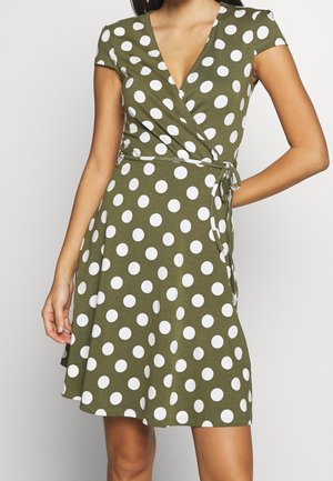 Kortærmet olivengrøn slå-om-kjole med store hvide polkaprikker, bundet i taljen, båret af en person med den ene hånd på hoften.