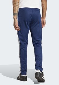 Námořnické sportovní kalhoty vyrobené z hladkého materiálu, s bílými bočními pruhy, zúženým střihem a zadní kapsou. Nosené s černými botami.