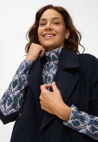 Cappotto di lana navy con rever a punta, indossato sopra un dolcevita a fantasia con motivi geometrici blu e verdi.