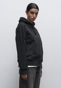 Felpa nera oversize con cappuccio e coulisse, polsini e orlo a coste. Indossata sopra una maglietta bianca, abbinata a pantaloni grigi. Texture piatta.