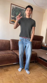 Jeune homme en chemise sombre et jean ample, debout pieds nus sur un parquet, tenant la main levée avec les doigts pincés, dans un salon avec un canapé marron.