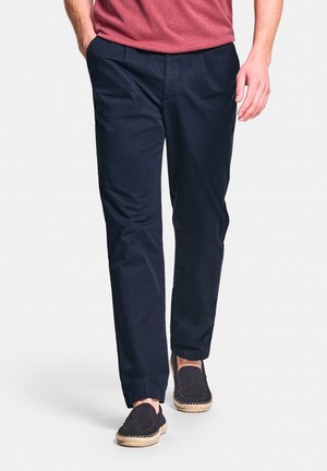 Man wearing navy blue pants, dark slip-on espadrilles with rope soles, and a red shirt, standing with one hand in pocket.