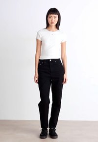 Sorte høye jeans med fem lommer, knappelukking og avslappet passform, parret med en hvit ribbestrikket t-skjorte med kort erm. Svarte joggesko på.