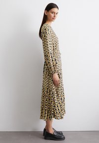 Langärmliges Kleid mit einem geometrischen Muster aus gelben, braunen und grauen Kreisen, mit einer taillierten Taille und einem fließenden Rock. Schwarze Plattformslipper.