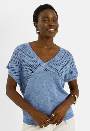 Mujer con un top de punto de manga corta azul claro y pendientes de aro dorados, pulseras y anillos, sonriendo con las manos entrelazadas delante.
