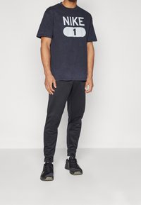 T-shirt en coton marine avec logo "NIKE" blanc et motif "1", associé à un pantalon de jogging noir et des chaussures de sport noires. Design simple, coupe décontractée.