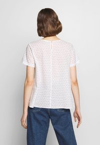 Biała bluzka z krótkim rękawem w kwiatowy wzór, z okrągłym dekoltem i małym guzikiem z tyłu. Wykonana z lekkiego materiału.