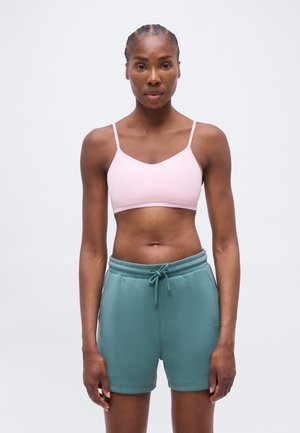 Atletisk kvinde, der står og har en lyserød sports-bh og turkis snøre-short på, mod en ensartet hvid baggrund.