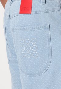Light blue denim shorts featuring a back pocket with white circular embroidery and a red leather brand tag at the waist.