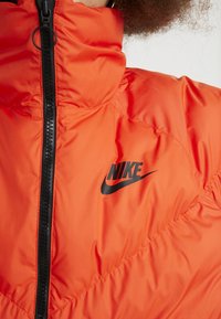 Nike puffer jacket in vibrant orange, featuring a high collar, black zipper, and prominent black logo on the chest. Smooth, padded texture.