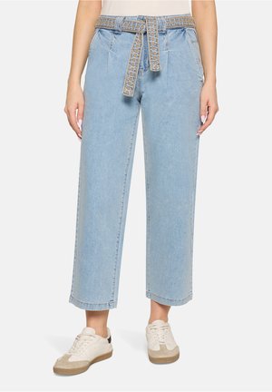 Kobieta w jasnoniebieskich szerokich dżinsach z wzorzystym paskiem z denimu zawiązanym w talii oraz białymi trampkami.