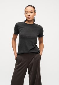 Νεαρή γυναίκα με πλεξούδες κοτσίδες, ντυμένη με εφαρμοστό μαύρο μπλουζάκι και φαρδιά σκούρα καφέ παντελόνια, στέκεται με τα χέρια στις τσέπες.