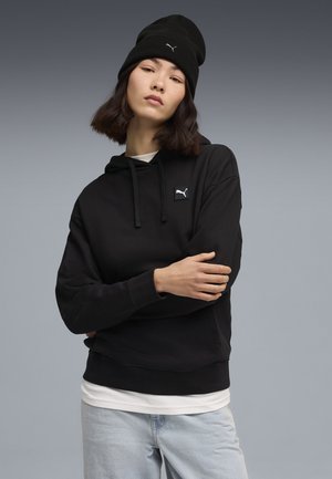Sort hættetrøje med snore og logo-lap på brystet, lagdelt over en hvid skjorte, parret med en sort beanie. Lyseblå jeans.