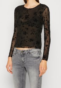 Svart mesh långärmad topp med blommönster och subtil glans, med rund halsringning och figurnära design. Matchad med grå denimjeans.