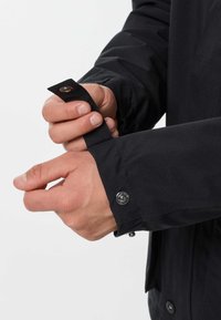 Mani che regolano il polsino con bottone a pressione di una giacca nera a maniche lunghe su uno sfondo chiaro e uniforme.