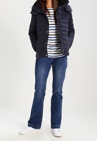 Osoba w granatowej kurtce puchowej, nosząca w paski koszulę, niebieskie dżinsy i białe trampki, stoi z rękami w kieszeniach kurtki.