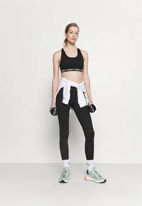 Črna športna modrček z logotipom na traku, črne legice in mint zelene športne superge. Bela jopa zavezana okoli pasu. Drži majhne uteži.