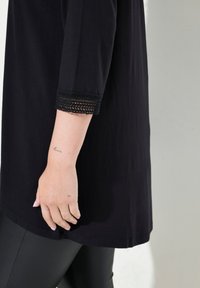 Zwarte tuniek met driekwart mouwen, voorzien van een kantafwerking bij de manchetten. Gecombineerd met zwarte, aansluitende legging. Gladde textuur.