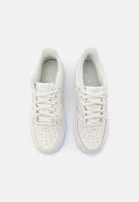 Baskets en cuir blanc à finition lisse, présentant des perforations sur le bout, des lacets plats et un logo Nike bleu clair sur le côté.