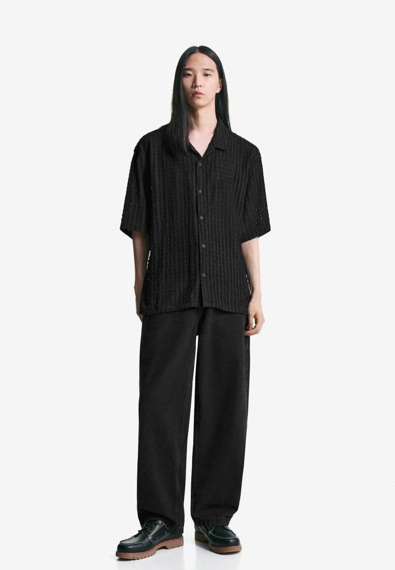Maglietta nera a maniche corte con un design a coste texturizzate, abbinata a pantaloni neri a gamba larga e scarpe nere con suola spessa.