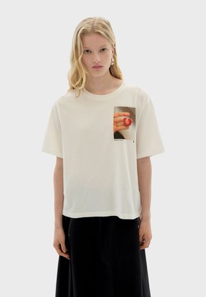 Witte katoenen t-shirt met korte mouwen, met een vierkant grafisch ontwerp van een hand die een hartvormig object vasthoudt. Ontspannen pasvorm en zachte textuur.