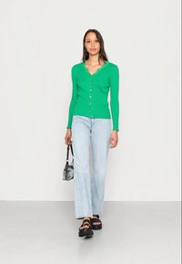 Cardigã verde tricotado com decote recortado, fecho de botão, combinado com jeans de perna reta azul claro e sandálias de plataforma pretas.