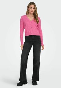 Cardigan rosa a coste con scollatura a V, chiusura con bottoni e maniche lunghe, abbinato a jeans neri a vita alta e stivali neri chunky.