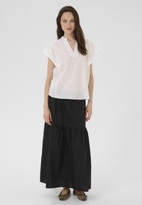 Blouse blanche à manches courtes avec décolleté en V et détails en œillets, associée à une longue jupe noire à volants, portée avec des ballerines marron.