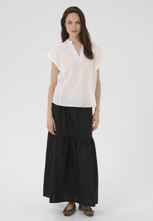 Blouse blanche à manches courtes avec décolleté en V et détails en œillets, associée à une longue jupe noire à volants, portée avec des ballerines marron.