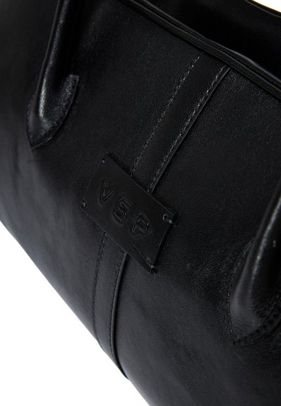 Primo piano di una borsa a tracolla in pelle nera con un'etichetta in pelle cucita, incisa con le lettere "VSP" sul pannello frontale.