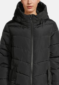 Frau trägt eine schwarze gesteppte Pufferjacke mit hohem Kragen und Reißverschluss, ausgestattet mit Kordelzügen an der Kapuze und Taschen mit Klappen.
