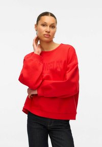 Rotes Sweatshirt mit übergroßen, puffigen Ärmeln und rundem Halsausschnitt. Mit einem auffälligen Textdesign auf der Vorderseite und einem lockeren Schnitt. Getragen mit dunklen Jeans.