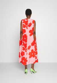 Vestido sin mangas de tela rosa con grandes patrones florales rojos, que cuenta con bolsillos laterales y una apertura en la parte posterior, combinado con tacones verdes.