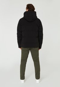 Chaqueta de plumas negra con capucha, textura acolchada y puños elásticos, combinada con pantalones verde oliva y zapatillas blancas, vista desde la parte trasera.