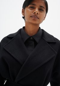 Manteau en mélange de laine noire avec un col en pointe et une silhouette structurée, présentant une finition texturée et une chemise noire à boutons en dessous.