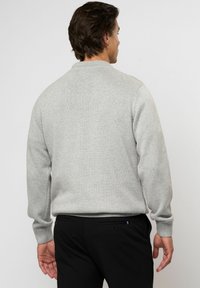 Muž s krátkymi hnedými vlasmi, oblečený v svetlosivom pletenom svetri a čiernych nohaviciach, stojaci chrbtom k divákovi na jednoduchom pozadí.