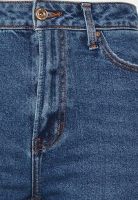 Jeans de mezclilla azul con una cintura alta, que presentan costuras naranjas, un cierre de botón metálico y una textura desvanecida en algunas áreas.