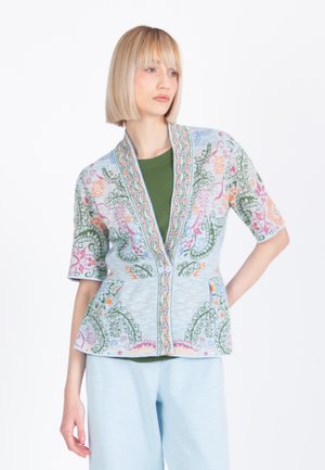 Mladá žena s krátkymi blond vlasmi má na sebe pastelový sveter s kvetinovým vzorom cez zelený top a svetlomodré nohavice, stojaca na bielom pozadí.