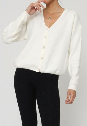 Cardigan tricoté blanc avec un col en V, des manches longues et six boutons à l'avant. Texture douce et coupe ample, associé à des leggings noirs.
