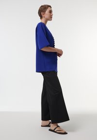 Camiseta azul oversized de manga corta, con un acento de volantes texturizado en la parte delantera, combinada con pantalones negros anchos y sandalias planas negras.