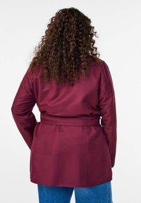 Burgunderrote taillierte Jacke mit glatter Textur, langen Ärmeln und einer maßgeschneiderten Passform, von hinten vor hellem Hintergrund gesehen.