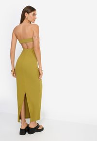 Μακρύ φόρεμα σε λαδί χρώμα χωρίς τιράντες με άνοιγμα στην πλάτη και σκίσιμο στο πλάι. Το μοντέλο φοράει μαύρα σανδάλια με πλατφόρμα και χρυσά αξεσουάρ.