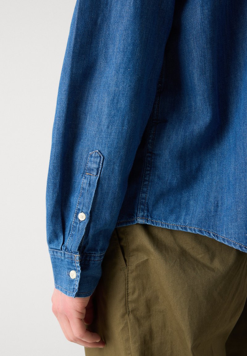 Dunkelblaue Jeanshemd mit langen Ärmeln, ausgestattet mit weißen Knöpfen und sichtbaren Nahtstichen. Kombiniert mit olivfarbenen Hosen.