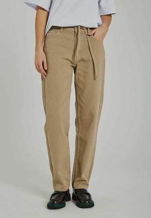Pantaloni beige a vita alta con taglio dritto, tasche anteriori e dettaglio con cordoncino in vita, abbinati a mocassini neri.